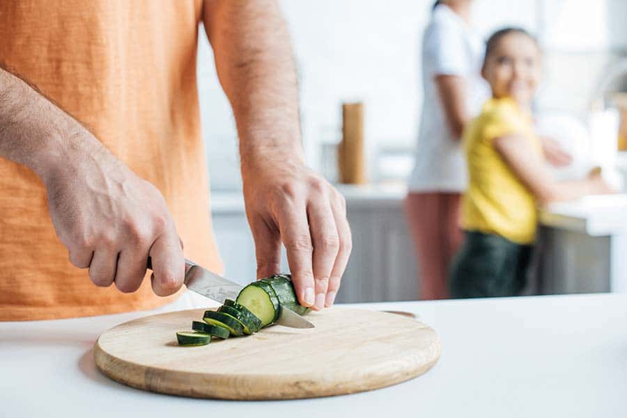 Мъж, който реже краставица на колелца в своята кухня. Назаден план неговото семейство също приготвя храна.