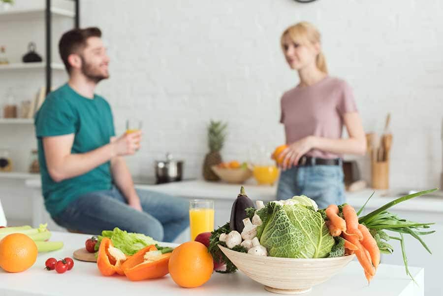 Млада двойка, хапваща плодове и зеленчуци в кухнята. На преден план са всички свежи зеленчуци.