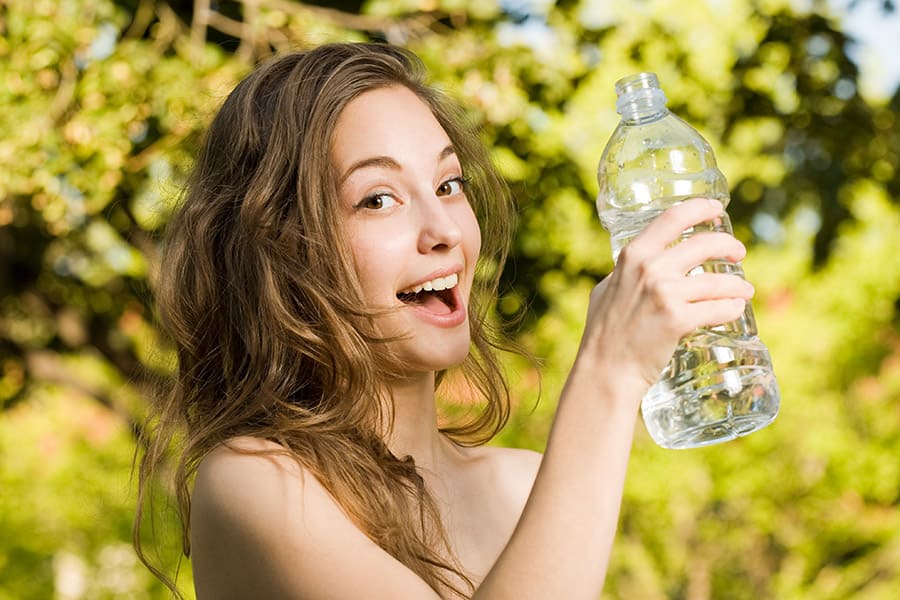 Млада жена сред природата държи в ръка пластмасова бутилка и се интересува от рисковете за здравето.