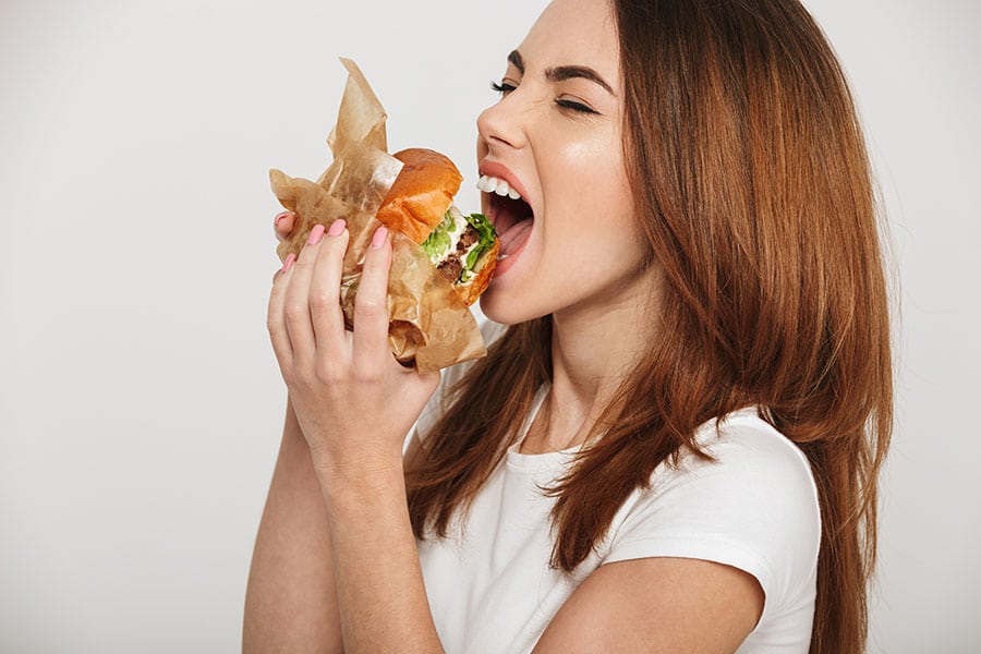 Вреди от въглехидратите. Млада жена яде хамбургер под въздействието на стреса.