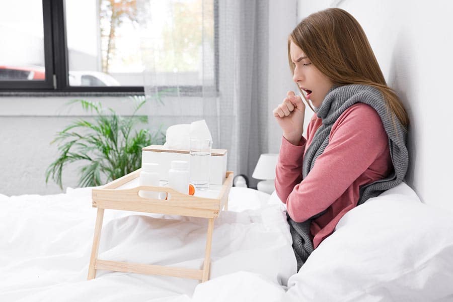 Жена, която кашля в леглото си с масичка, върху която има чаша с билки при респиратоени инфекции и проблеми с дихателна система